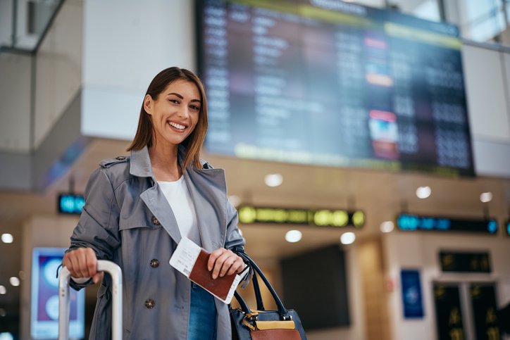 Happy woman in front of information board at airport terminal looking at camera.